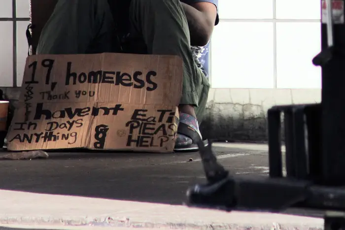 A homeless teenager sits on a sidewalk with a cardboard sign saying he's 19 and hasn't eaten